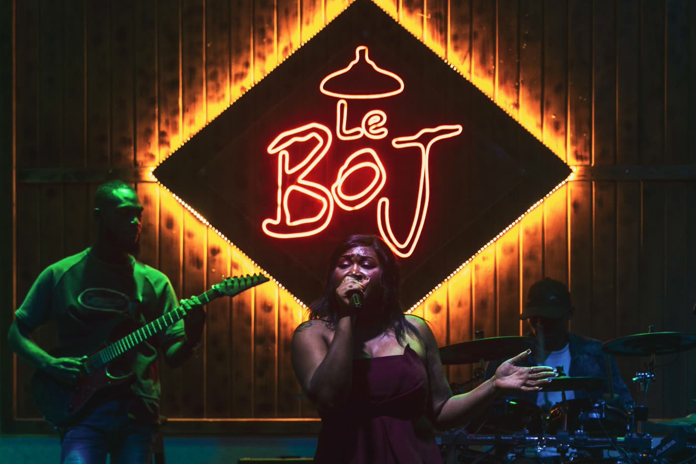 Chanteuse au micro, guitariste et batteur sur la scène du Boj, sous l'enseigne néon